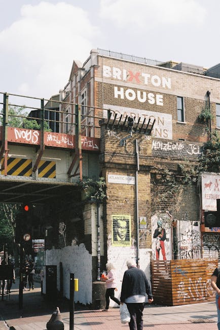 View of the exterior of a brick building at Brixton Market in Lambeth, with large painted signs reading 'BRIXTON HOUSE' on the front facade. The building features an elevated platform with a metal railing, scaffolding-like structure, and graffiti-covered walls. In the foreground, pedestrians are walking along the pavement, with some entering or exiting a covered passageway beneath the building. Part of the scene shows a wooden pallet, possibly used in furniture transport or packaging. The lighting is natural, with a partly cloudy sky overhead. This setting relates to home relocation and furniture transport services provided by Lambeth Removals, highlighting the urban environment characteristic of house removals within the area.