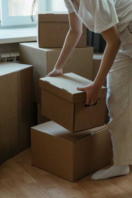 A person, dressed in a light-colored t-shirt and white pants, is seen organizing cardboard moving boxes inside a room with natural light coming through a window. The boxes vary in size, with some labeled, such as one marked 'CLOTHES', and are stacked on top of each other or being prepared for loading. The individual is in the process of lifting or positioning a box, using both hands for stability. The setting suggests an interior space during a home relocation, with wooden flooring visible and additional boxes and packaging materials nearby. This scene illustrates the packing stage of a furniture transport or house removals process, common to services provided by Lambeth Removals, focusing on careful handling and logistics involved in loading goods for a seamless move.