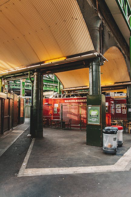 View of the exterior of a brick building at Brixton Market in Lambeth, with large painted signs reading 'BRIXTON HOUSE' on the front facade. The building features an elevated platform with a metal railing, scaffolding-like structure, and graffiti-covered walls. In the foreground, pedestrians are walking along the pavement, with some entering or exiting a covered passageway beneath the building. Part of the scene shows a wooden pallet, possibly used in furniture transport or packaging. The lighting is natural, with a partly cloudy sky overhead. This setting relates to home relocation and furniture transport services provided by Lambeth Removals, highlighting the urban environment characteristic of house removals within the area.