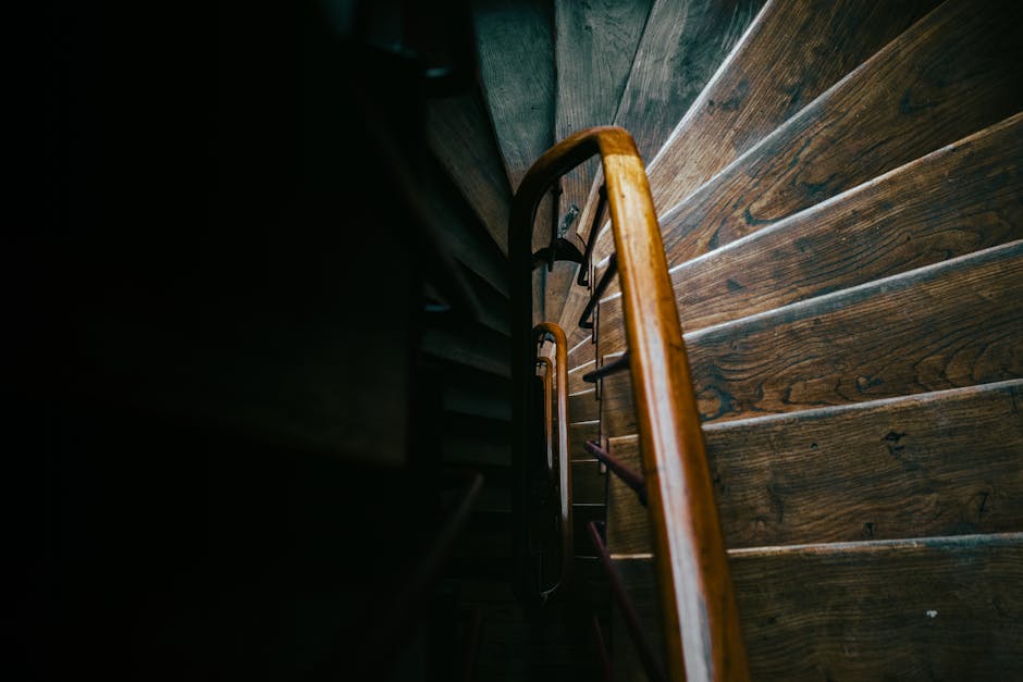 A narrow wooden staircase viewed from above, displaying dark, polished wooden steps and a curved wooden handrail. The staircase is inside a property, with a portion of the wall visible on the left side, painted in a dark colour. The steps are closely spaced, creating a confined ascending space, while the lighting emphasizes the natural grain of the wood. In the background, the staircase continues upwards, with a landing partially visible at the top. This image captures the typical challenge of navigating narrow staircases during house removals or moving jobs, which Lambeth Removals often encounters when handling home relocation or furniture transport within tight interior spaces. The environment appears well-maintained, but the limited width of the stairs highlights potential difficulties in moving large or bulky items, requiring careful planning and equipment such as trolleys or packing blankets to facilitate safe transport. The scene underscores the importance of professional assistance in managing the complexities of packing and moving in such constrained environments.