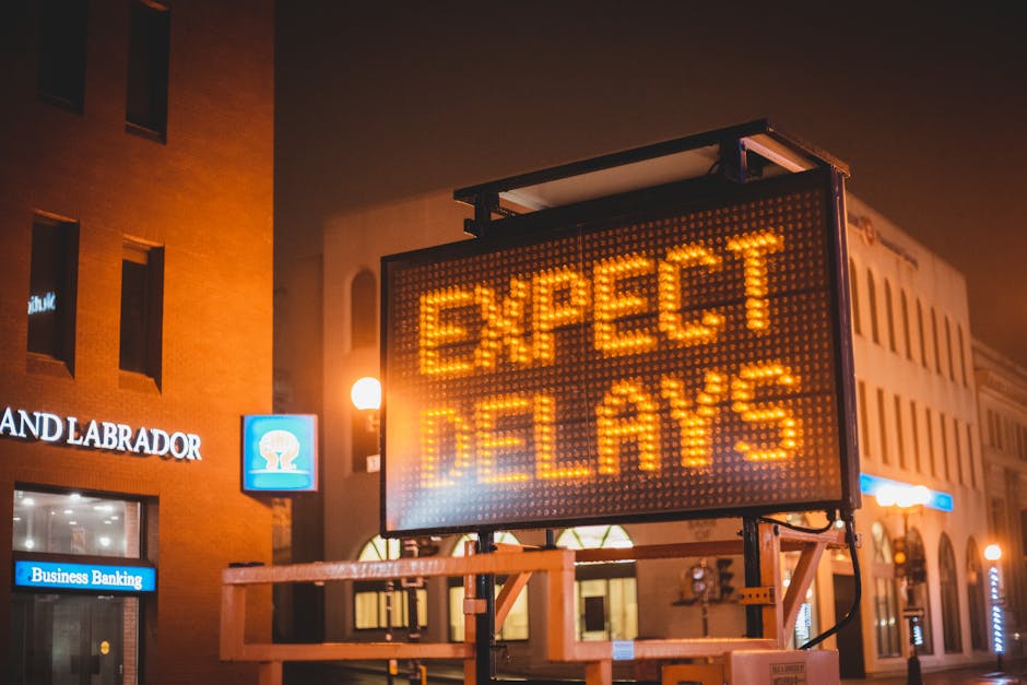 A large illuminated electronic road sign displaying the message 'EXPECT DELAYS' in bright orange lights is positioned on a metal stand on a city street at night. The sign is mounted on a wooden platfo