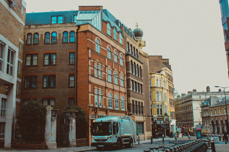 A city street scene showing a Lambeth Removals vehicle parked outside multi-storey buildings with a mix of brick and ornate architectural details, including arched windows and decorative facades. The vehicle, a white van with signage for furniture transport services, is positioned on the pavement near the entrance of an urban property, suggesting a home relocation or moving process. Several cardboard boxes and wrapped furniture items are visible on the sidewalk, ready for loading or unpacking. A trolley and straps are nearby, indicating active handling of household items. The background features additional buildings with classic architectural elements, street lamps, and bicycles locked along the sidewalk. Bright daylight illuminates the scene, highlighting the typical busy atmosphere of a London neighbourhood involved in packing and moving activities, supported by Lambeth Removals' professional services during furniture transport and house removals.