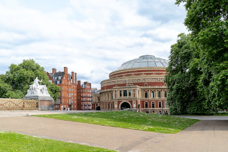 A view of the Royal Albert Hall, a large circular Victorian concert hall with a prominent domed roof made of grey metal, situated in a park area with well-maintained green grass, walking paths, and mature trees. To the left of the hall, there is a white stone sculpture depicting a seated figure surrounded by other figures, located on a pedestal. Behind the sculpture, a barricade made of metal fencing is visible. Multi-storey red brick buildings with traditional architecture are seen in the background behind the hall and sculpture. The sky above is partly cloudy with patches of blue sky, and the scene appears to be daytime. The overall setting reflects a cultural and historical landscape, potentially relevant to house removals or furniture transport services in central London, such as those provided by Lambeth Removals.