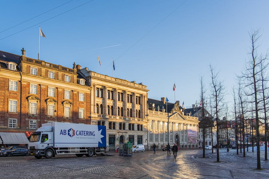A city street scene showing a Lambeth Removals vehicle parked outside multi-storey buildings with a mix of brick and ornate architectural details, including arched windows and decorative facades. The vehicle, a white van with signage for furniture transport services, is positioned on the pavement near the entrance of an urban property, suggesting a home relocation or moving process. Several cardboard boxes and wrapped furniture items are visible on the sidewalk, ready for loading or unpacking. A trolley and straps are nearby, indicating active handling of household items. The background features additional buildings with classic architectural elements, street lamps, and bicycles locked along the sidewalk. Bright daylight illuminates the scene, highlighting the typical busy atmosphere of a London neighbourhood involved in packing and moving activities, supported by Lambeth Removals' professional services during furniture transport and house removals.
