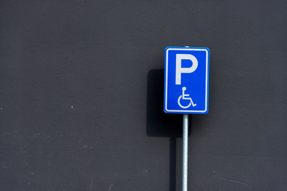 A blue rectangular parking sign with a white letter 'P' and a white wheelchair symbol indicating accessible parking, mounted on a metallic pole positioned against a dark grey wall. The sign casts a shadow on the wall, highlighting the textured surface. This image relates to parking regulations for house removals and relocation services, showcasing the importance of designated accessible parking spaces, which Lambeth Removals may need to consider during street parking and loading processes for home relocation and furniture transport. The setting suggests an outdoor urban environment, suitable for documenting permit rules for street parking during move times as specified by Lambeth Council, with the sign serving as a visual cue for parking compliance in moving logistics.