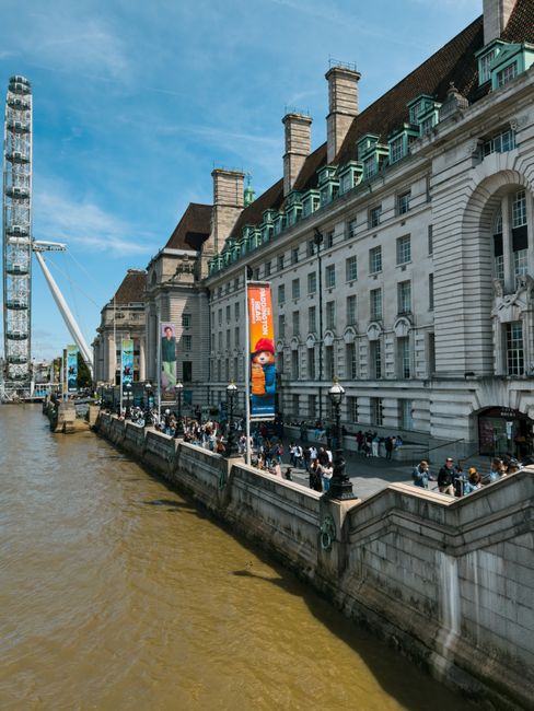 A wide view of the London skyline during daytime, featuring a river in the foreground with several docked boats and a bridge crossing over the water. On the left, modern office and residential buildin
