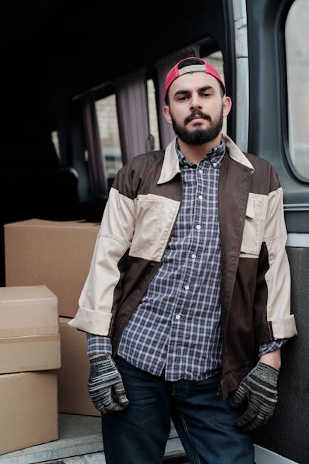 A young man with a beard and wearing a red cap, a checked shirt, and a beige and brown jacket stands inside a van used for home relocation and furniture transport. The interior of the van is visible, showing several cardboard boxes stacked behind him, some wrapped in plastic or fabric for protection during moving. The man is wearing gloves, indicating he is involved in the packing and loading process. Light enters through the van's windows, illuminating the scene, and the vehicle appears to be on a street or driveway, ready for or in the process of a house removal. Lambeth Removals, a professional moving company, provides this service as part of their comprehensive removals category, supporting efficient and careful transportation of household items within the Brixton and Lambeth area.