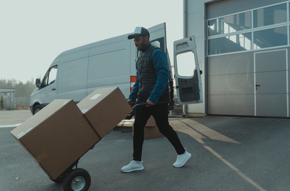 A young man with a beard and wearing a red cap, a checked shirt, and a beige and brown jacket stands inside a van used for home relocation and furniture transport. The interior of the van is visible, showing several cardboard boxes stacked behind him, some wrapped in plastic or fabric for protection during moving. The man is wearing gloves, indicating he is involved in the packing and loading process. Light enters through the van's windows, illuminating the scene, and the vehicle appears to be on a street or driveway, ready for or in the process of a house removal. Lambeth Removals, a professional moving company, provides this service as part of their comprehensive removals category, supporting efficient and careful transportation of household items within the Brixton and Lambeth area.