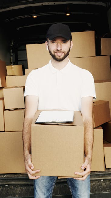 A young man with a beard, wearing a black cap and a white polo shirt, is standing inside the open back of a moving van, holding a medium-sized cardboard box. The van's interior is visible, filled with stacked cardboard boxes of various sizes, some covered with plastic wrap. The man is smiling slightly and appears to be in the process of loading or unloading boxes as part of a house removal. The scene is outdoors, with natural light illuminating the area, and the surrounding environment suggests a residential or urban setting. This image illustrates the packing and furniture transport aspects of home relocation, relevant to Lambeth Removals' services for last-minute or urgent same-day moves.