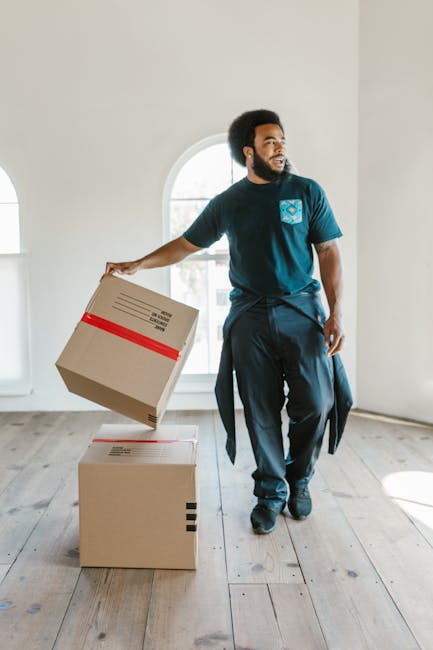 A man with dark curly hair and a beard, wearing a blue T-shirt with a protective packing logo and dark trousers, is inside a bright, spacious room with white walls and wooden flooring. He is in the process of moving, holding a cardboard box with red and black packing tape in his right hand, while two additional cardboard boxes, sealed with packing tape and labels, are positioned on the floor nearby. The background features a large arched window with natural daylight illuminating the space, indicating a home environment. This scene depicts the packing and loading process associated with house removals, with the man preparing items for transport, possibly for a home relocation or furniture transport service, as part of Lambeth Removals’ ongoing moving logistics.