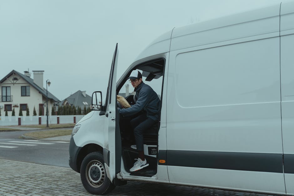 A young man with a beard, wearing a black cap and a white polo shirt, is standing inside the open back of a moving van, holding a medium-sized cardboard box. The van's interior is visible, filled with stacked cardboard boxes of various sizes, some covered with plastic wrap. The man is smiling slightly and appears to be in the process of loading or unloading boxes as part of a house removal. The scene is outdoors, with natural light illuminating the area, and the surrounding environment suggests a residential or urban setting. This image illustrates the packing and furniture transport aspects of home relocation, relevant to Lambeth Removals' services for last-minute or urgent same-day moves.