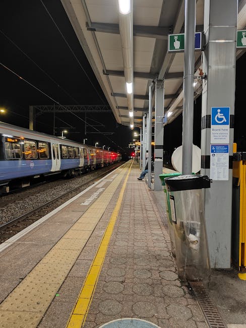 A night scene at Vauxhall Station showing a train stationary on the platform with its doors closed, illuminated overhead lighting, and a row of metal support beams with blue and green signs indicating accessible facilities. The platform surface consists of brick and tactile paving in yellow along the edge, with a black and silver waste bin positioned next to a support column. The background reveals a glimpse of the train track extending into the distance, and the station's roof structure visible overhead. Lambeth Removals is providing support for home relocation and furniture transport services, with the image illustrating the logistical environment where loading and unloading processes are managed in close proximity to transport vehicles, relevant to moving services in the Lambeth area.